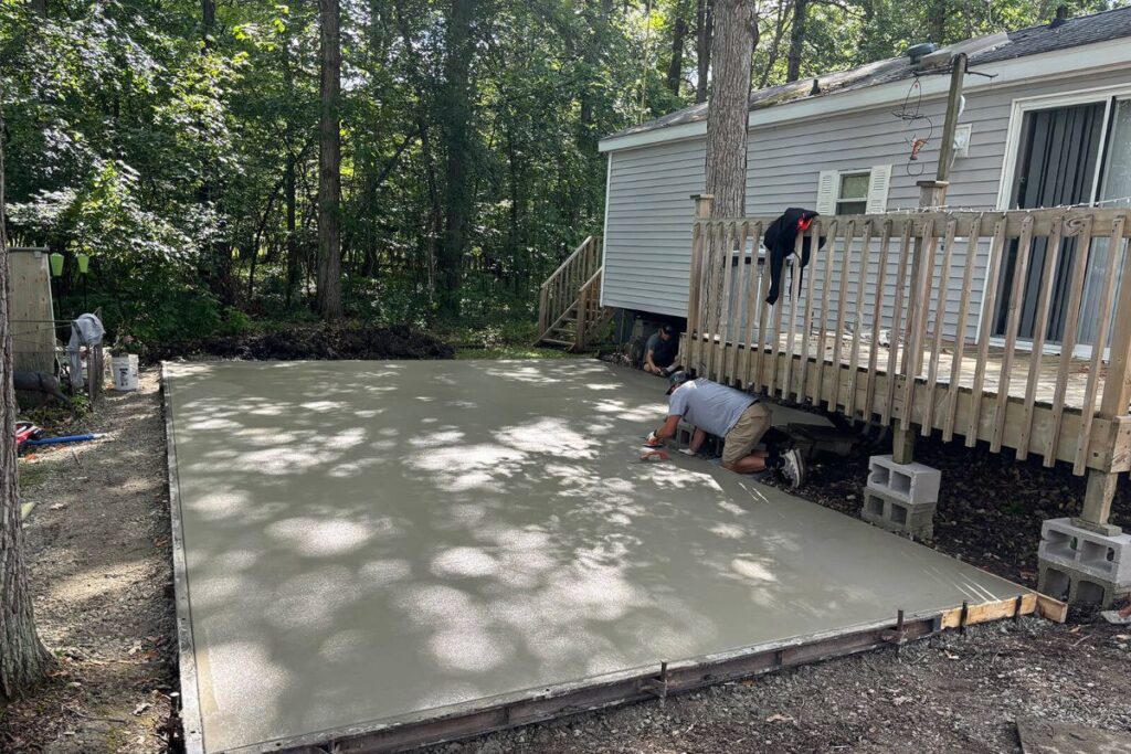 Crew members from Concrete Landscape Solutions pouring and leveling a new concrete patio extension next to a wooden deck at a home in Riverbend RV Park, Watertown, WI.