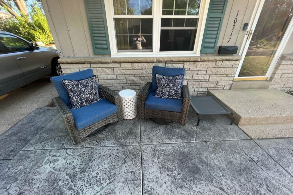 A close-up view of the new dark gray stamped concrete front patio, showing the intricate texture and pattern with two chairs and a side table.